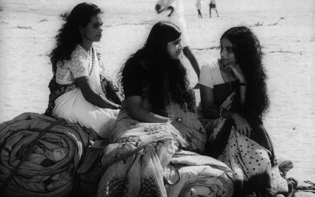 Filmstill aus THAMP: Drei in Saris gekleidete Frauen sitzen auf zu Bündeln zusammengerollten Decken auf dem Boden.