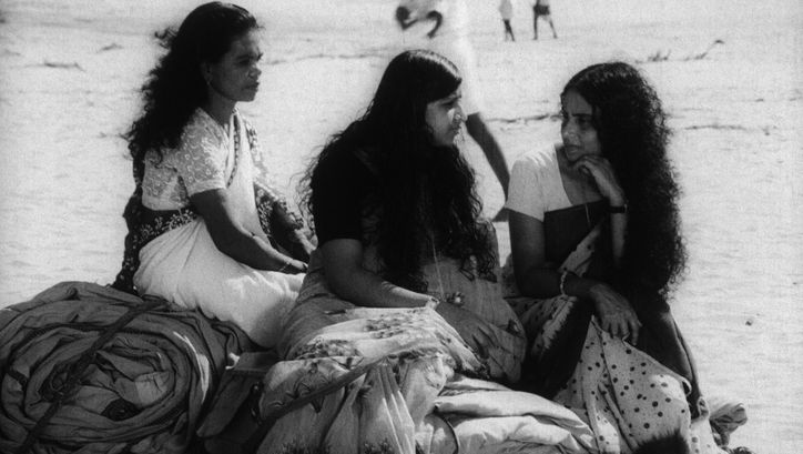 Film still from THAMP: Three women dressed in saris are sitting on rolled up blankets on the floor.