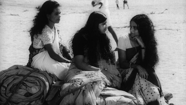 Filmstill aus THAMP: Drei in Saris gekleidete Frauen sitzen auf zu Bündeln zusammengerollten Decken auf dem Boden.