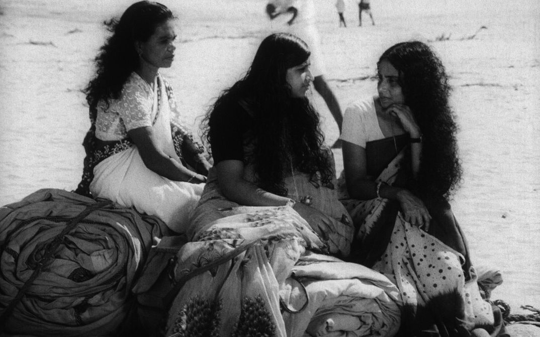 Film still from THAMP: Three women dressed in saris are sitting on rolled up blankets on the floor.