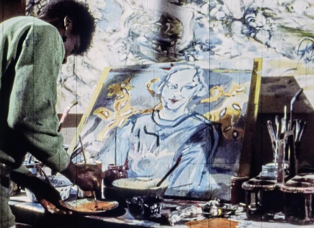 Filmstill aus „A Lover & Killer of Colour“ von Wanjiru Kinyanjui. Eine Frau mischt Farben mit einem Pinsel auf einem Teller. Auf einem Tisch stehen eine bemalte Leinwand und Schüsseln mit Pinseln und Farben.