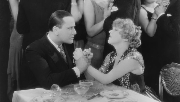 Film still from CHAMPAGNE: An elegantly dressed couple at a table, they look emotional. He holds her hands tightly. Couples dancing in the background.