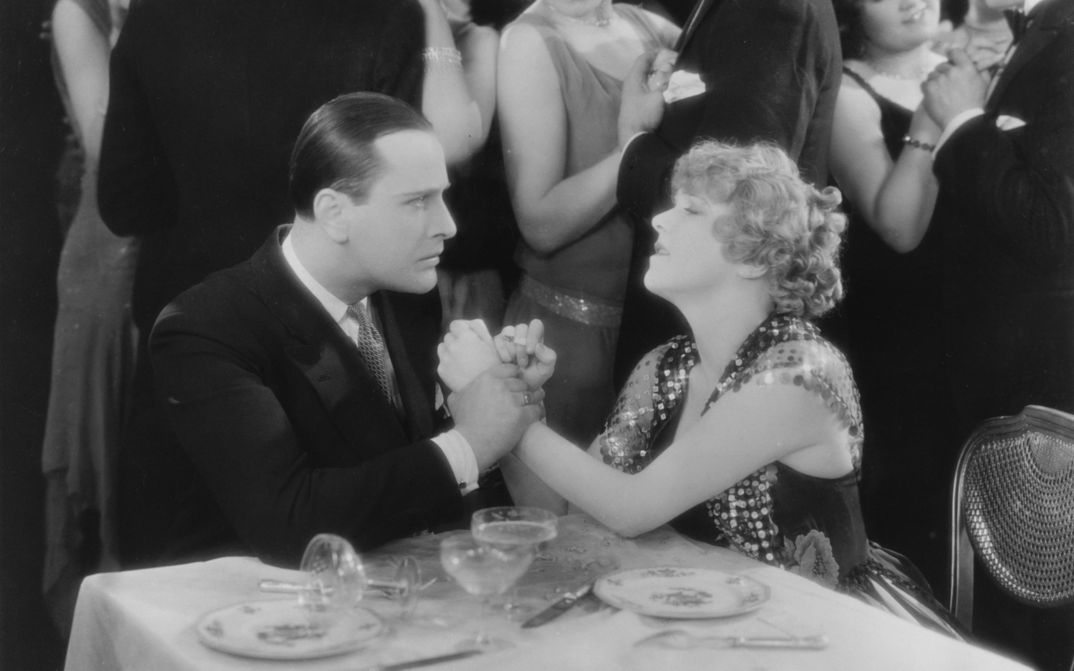 Filmstill aus CHAMPAGNE: Ein elegant gekleidetes Paar an einem Tisch, sie wirken emotional. Er hält ihre Hände fest. Im Hintergrund tanzende Paare.