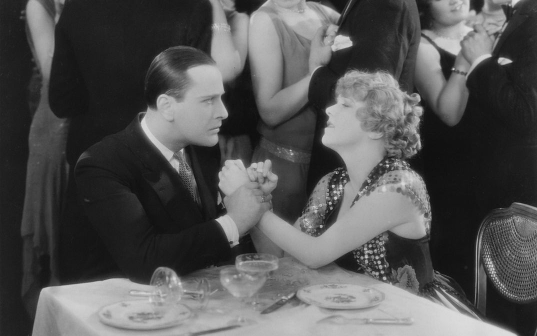 Film still from CHAMPAGNE: An elegantly dressed couple at a table, they look emotional. He holds her hands tightly. Couples dancing in the background.
