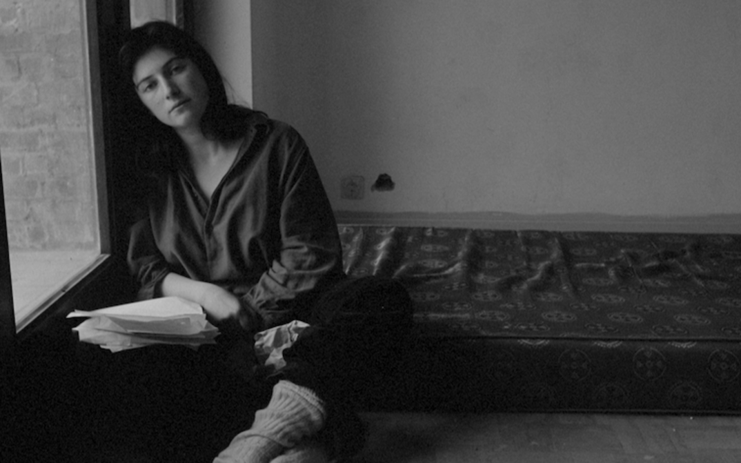 Filmstill aus JE, TU, Il, ELLE: Eine junge Frau mit langen Haaren sitzt in einem Zimmer am Boden und lehnt sich an die Wand. Auf ihrem Schoß liegen mehrere Zettel, im Hintergrund ist eine Matratze zu sehen.