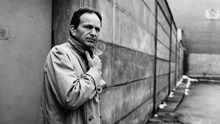 Filmstill aus DER TANGOSPIELER: Ein Mann steht vor einer Mauer, er hält den Kragen seines Mantels zu.