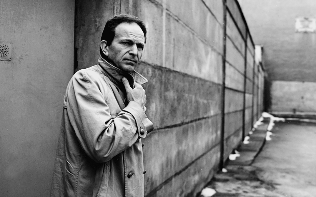 Film still from DER TANGOSPIELER: A man stands in front of a wall, holding the collar of his coat closed.