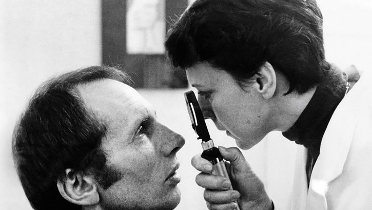 Black and white image of a middle-aged man and a female doctor, both are in profile to the camera. The doctor is looking slightly down at the sitting man, studying his eye with a magnifying lens.