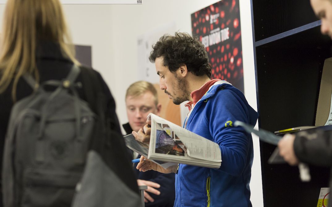 In einem Raum stehen ein paar Menschen. In der Mitte steht ein Mann, der aufmerksam ein Magazin liest.