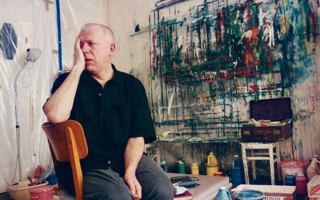 Künstler sitzt im Atelier vor bemalter Wand, Hand im Gesicht.