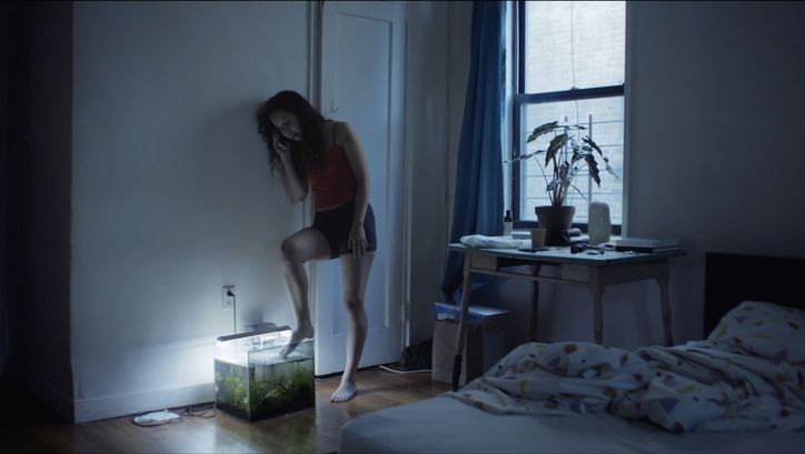 Film still from ACTUAL PEOPLE. A young woman in shorts and top leans against the wall in a bedroom, making a phone call. She holds her toes in an aquarium standing on the floor. 