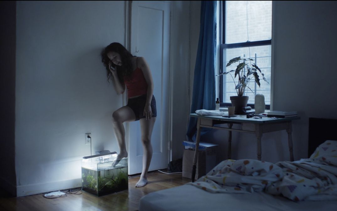 Film still from ACTUAL PEOPLE. A young woman in shorts and top leans against the wall in a bedroom, making a phone call. She holds her toes in an aquarium standing on the floor. 