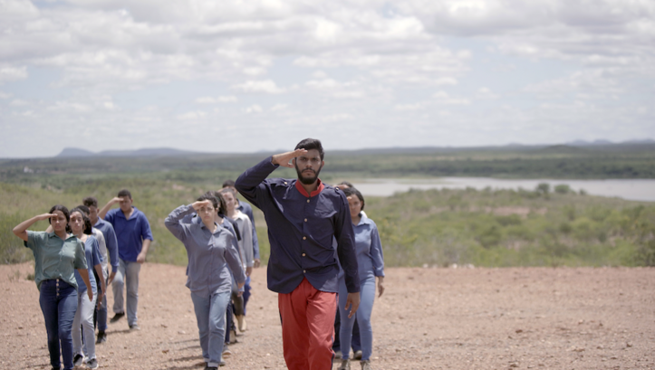 Film still from Cuitá, Stone From The Sky: A man in uniform leads a group of people in civilian clothing in marching.