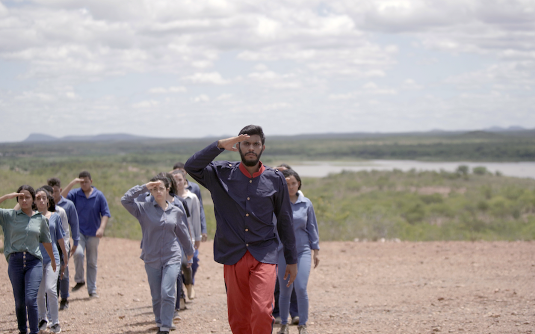 Film still from Cuitá, Stone From The Sky: A man in uniform leads a group of people in civilian clothing in marching.