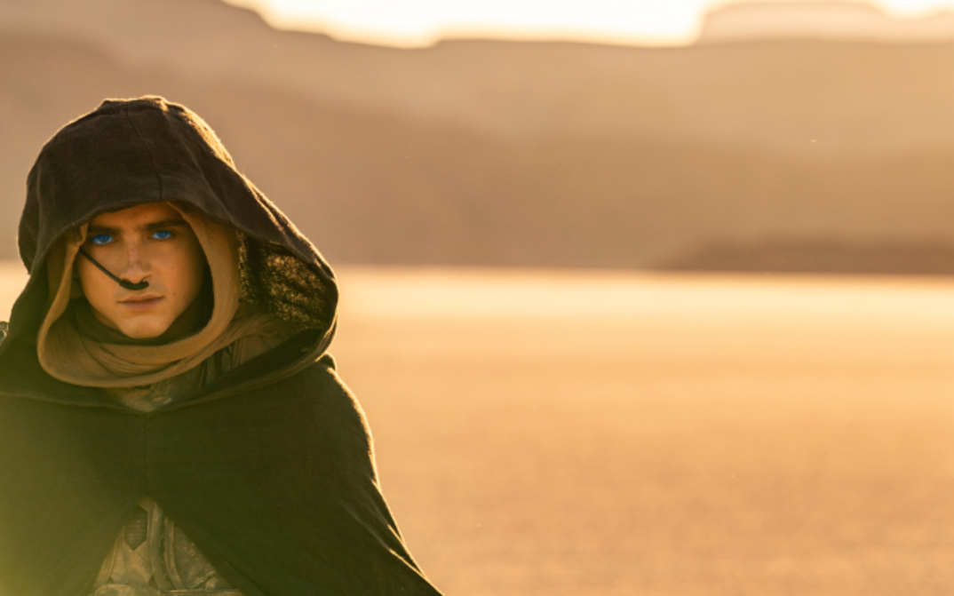 Filmstill from DUNE 2: A young man in the desert. He is wearing a cloak with a hood, has very blue eyes and a tube on his nose.