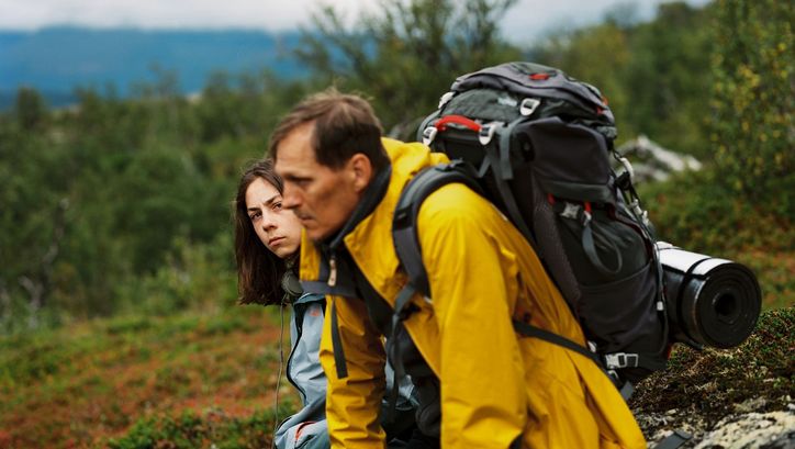 Filmstill from HELLE NÄCHTE: A man with a hiking rucksack and sleeping mat and another younger person are sitting on a rock in the mountains. Trees and rocks can be seen.