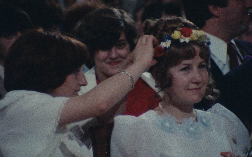 Filmstill aus LA ROSIÈRE DE PESSAC 79: Zwei Frauen machen einen Kranz in das Haar eines jungen, festlich gekleideten Mädchens.