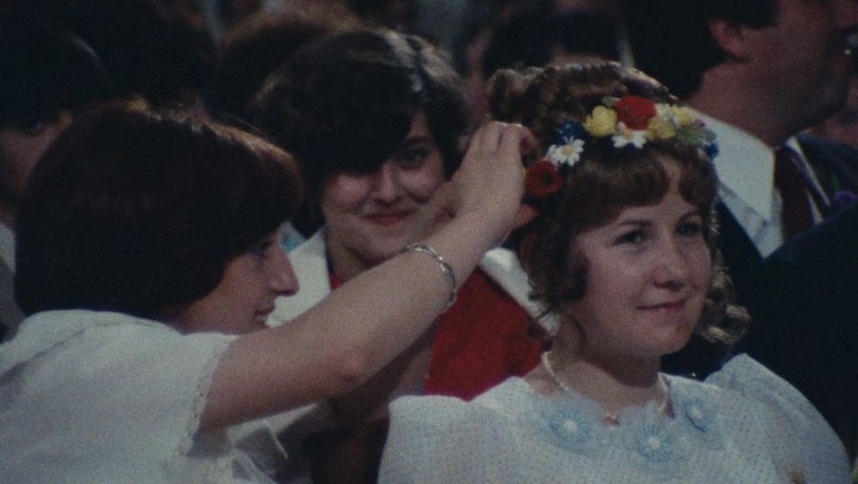 Filmstill aus LA ROSIÈRE DE PESSAC 79: Zwei Frauen machen einen Kranz in das Haar eines jungen, festlich gekleideten Mädchens.