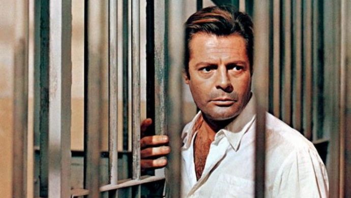 Film still from LO STRANIERO: Marcello Mastroianni is seen between the bars of a prison.