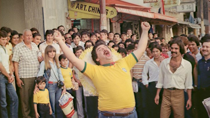 Filmstill aus NOITE PARAGUAYAS: Ein Mann in einem gelben T-Shirt steht inmitten einer Menschenmenge auf der Straße und streckt vor Freude die Arme in die Höhe.