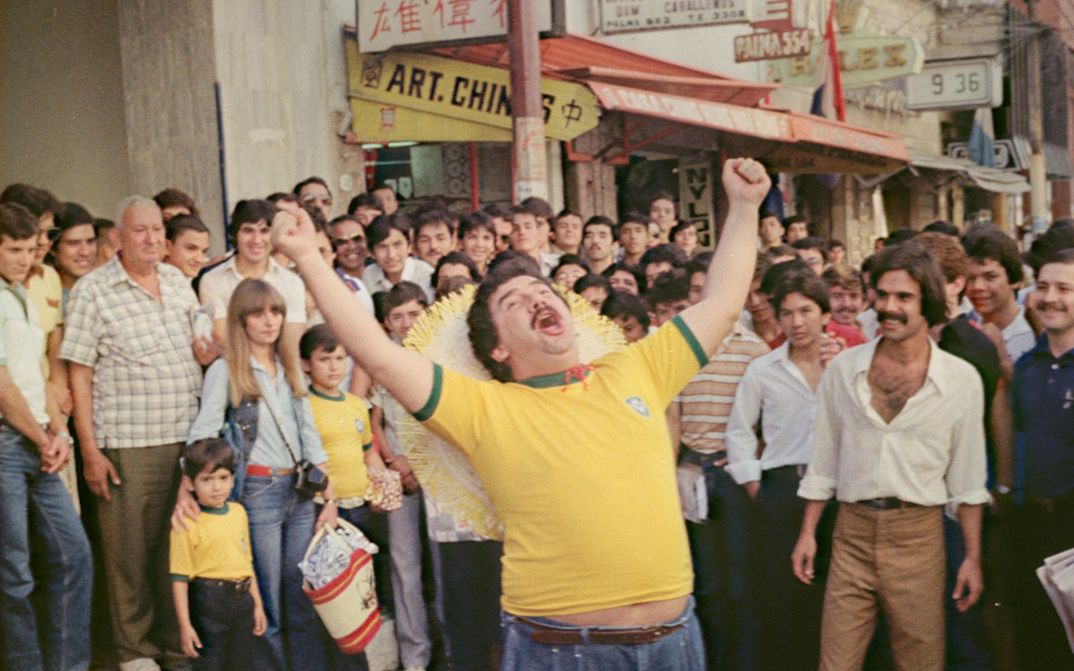 Filmstill aus NOITE PARAGUAYAS: Ein Mann in einem gelben T-Shirt steht inmitten einer Menschenmenge auf der Straße und streckt vor Freude die Arme in die Höhe.