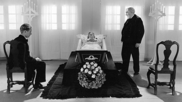 Film still from ORDET: In the center of the picture in black and white, a woman lies in a coffin. Two men are looking at her on the right and left. One is young, one is old, both are wearing dark suits. 