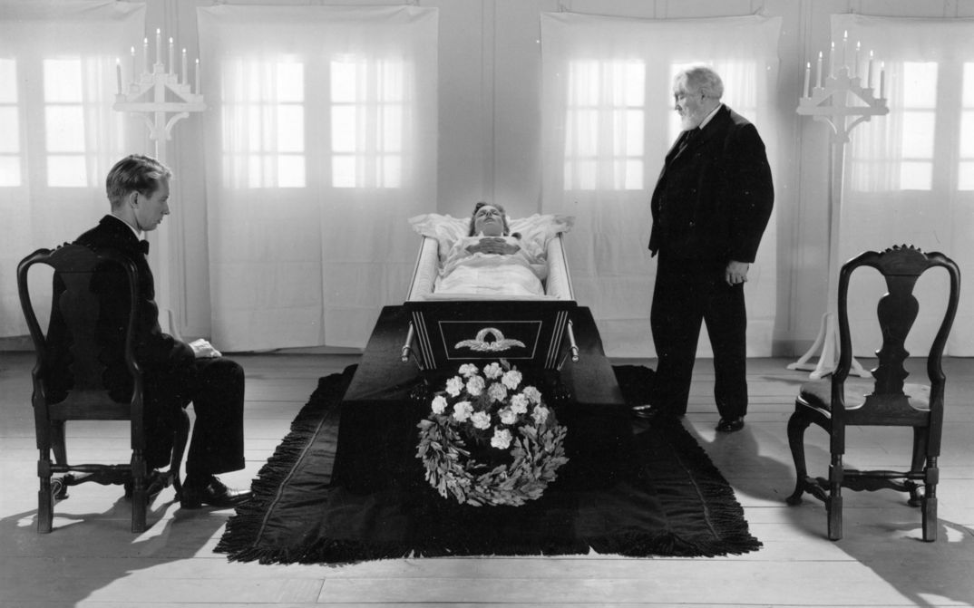 Film still from ORDET: In the center of the picture in black and white, a woman lies in a coffin. Two men are looking at her on the right and left. One is young, one is old, both are wearing dark suits. 