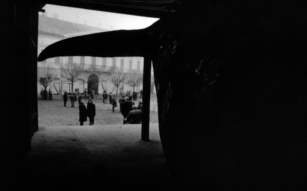 Filmstill aus WERCKMEISTER HARMÓNIÁK: Aus einem dunklen Raum heraus, in dem man die Schwanzflosse des ausgestopften Wales sieht, blickt man auf einen Platz mit winterlich gekleideten Menschen.
