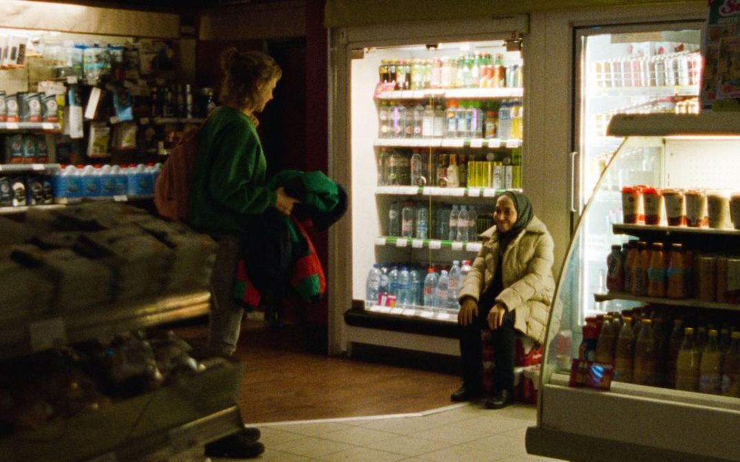 Filmstill aus GHOST TROPIC: In einem schwach beleuchteten Supermarkt sind zwei Frauen zu sehen, die sich anlächeln.