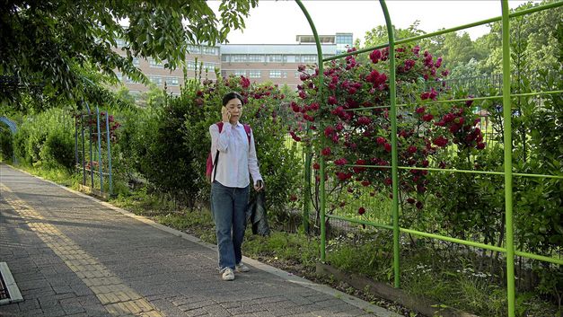 Still from the film "Hot in Day, Cold at Night" by Park Song-yeol. A woman speak into a cell phone as she walks on a bike path, next in front of a fence covered in shrubs.