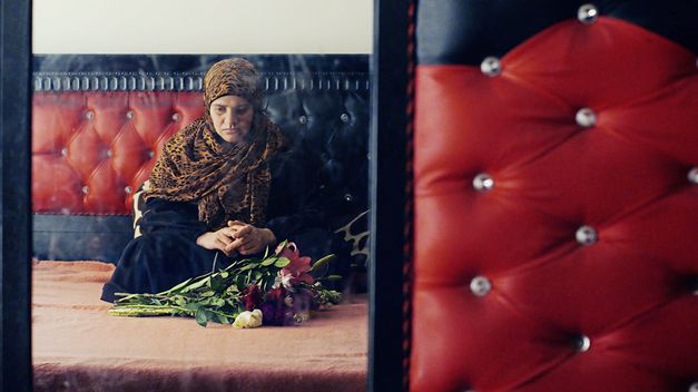 Filmstill aus „Anqa" von Helin Çelik. Eine verschleierte Frau sitzt meditativ vor einem Blumenstrauß.