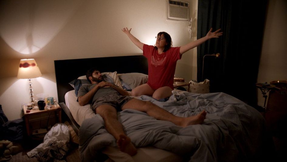 Filmstill aus THE FEELING THAT THE TIME FOR DOING SOMETHING HAS PASSED: Ein Mann und eine Frau in einem ungemachen Bett. Beide tragen eine Unterhose und T-Shirt. Sie sitzt und hält die Arme ausgestreckt.