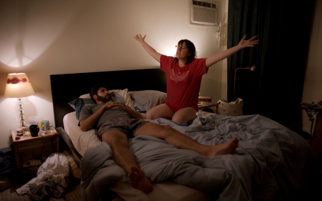 Filmstill aus THE FEELING THAT THE TIME FOR DOING SOMETHING HAS PASSED: Ein Mann und eine Frau in einem ungemachen Bett. Beide tragen eine Unterhose und T-Shirt. Sie sitzt und hält die Arme ausgestreckt.