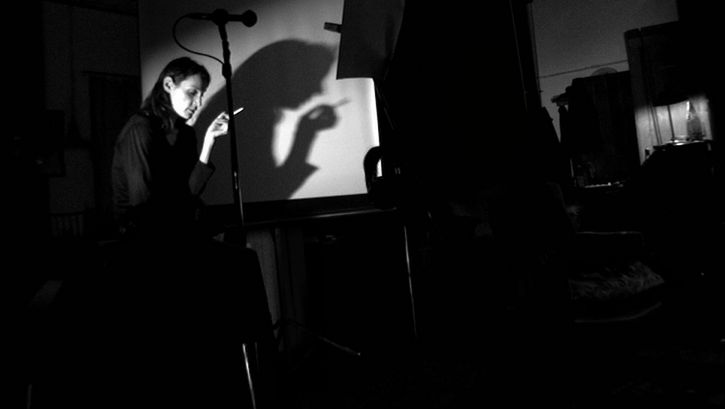 Filmstill aus NE CHANGE RIEN: In einem dunklen Raum steht eine Frau mit einer Zigarette in der Hand vor einer Leinwand. Vor ihr stehen ein Mikrophon und ein Notenpult.