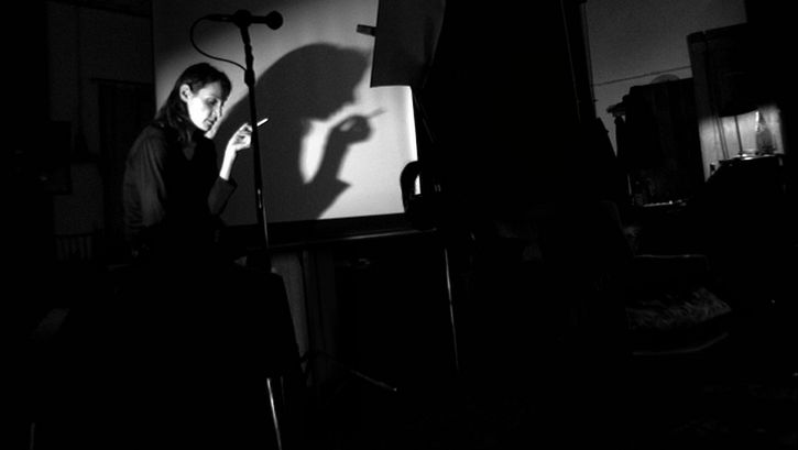 Film still from NE CHANGE RIEN: In a dark room, a woman stands in front of a screen with a cigarette in her hand. In front of her are a microphone and a music stand.
