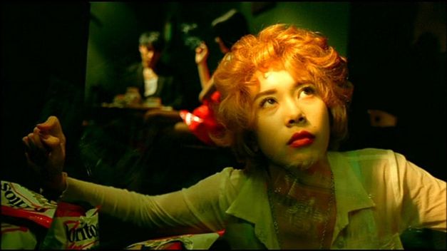 Filmstill aus FALLEN ANGELS: Nahaufnahme einer jungen Frau mit rötlich-blond gefärbten Locken. Sie blickt nach oben, hinter ihr sind Menschen in einem Lokal zu sehen. 