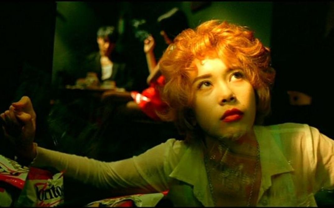 Filmstill aus FALLEN ANGELS: Nahaufnahme einer jungen Frau mit rötlich-blond gefärbten Locken. Sie blickt nach oben, hinter ihr sind Menschen in einem Lokal zu sehen. 