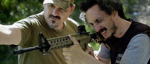 Still from the film "Camuflaje (Camouflage)" by Jonathan Perel. Two men in medium-close up look down the barrel of an automatic rifle. 
