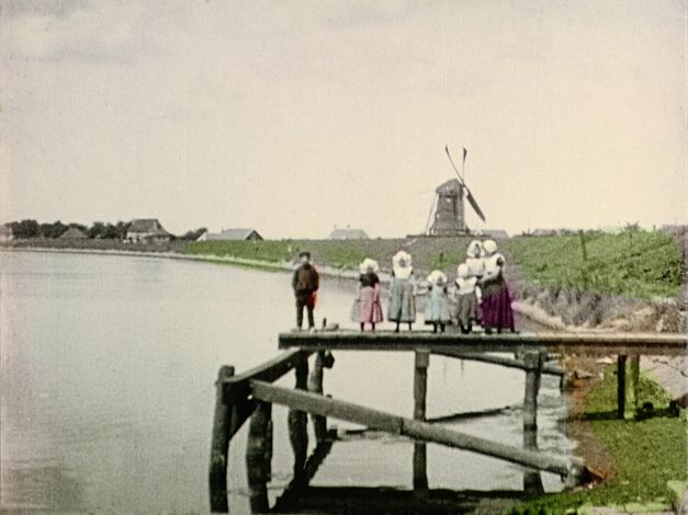 Filmstill from "Dearest Fiona" by Fiona Tan. Old image of a family on a wooden pontoon. In the distance, we see a mill and some houses. The clothes, the vegetation and the water are hand colored. The rest is in black and white.