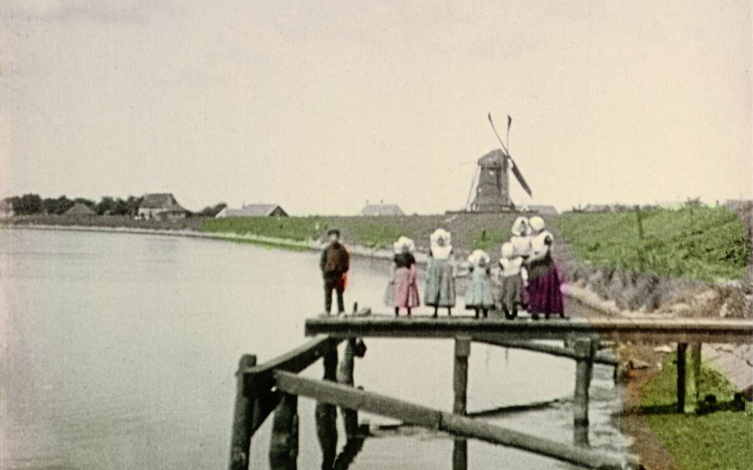 Filmstill from "Dearest Fiona" by Fiona Tan. Old image of a family on a wooden pontoon. In the distance, we see a mill and some houses. The clothes, the vegetation and the water are hand colored. The rest is in black and white.