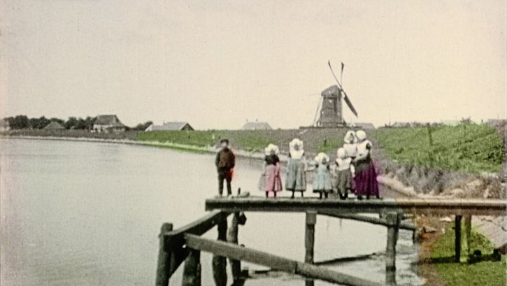 Filmstill from "Dearest Fiona" by Fiona Tan. Old image of a family on a wooden pontoon. In the distance, we see a mill and some houses. The clothes, the vegetation and the water are hand colored. The rest is in black and white.