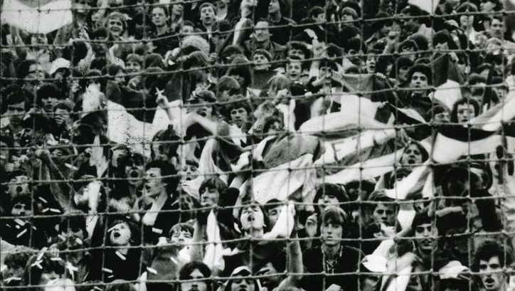 Filmstill aus ARÉNA. In schwarz-weiß sieht man jubelnde Zuschauer*innen eines Sportanlasses, sie sind hinter einem Zaun.