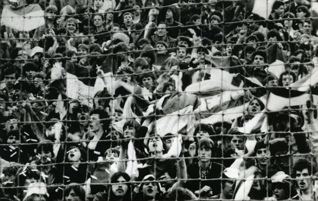 Filmstill aus ARÉNA. In schwarz-weiß sieht man jubelnde Zuschauer*innen eines Sportanlasses, sie sind hinter einem Zaun.