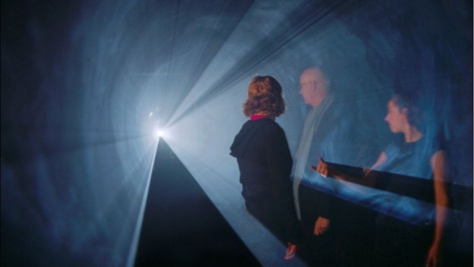 Film still from LINE DESCRIBING A CONE: Three people are standing in a cone of light in an otherwise dark room.