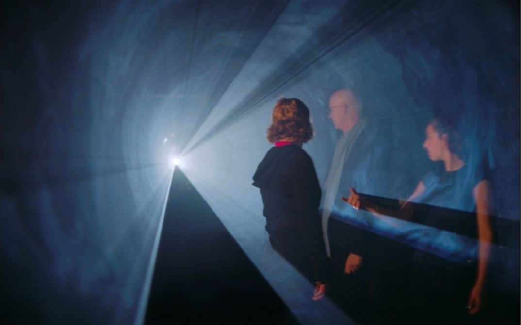 Film still from LINE DESCRIBING A CONE: Three people are standing in a cone of light in an otherwise dark room.