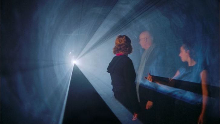 Filmstill aus LINE DESCRIBING A CONE: Drei Personen stehen in einem Lichtkegel in einem ansonsten schwarzen Raum.