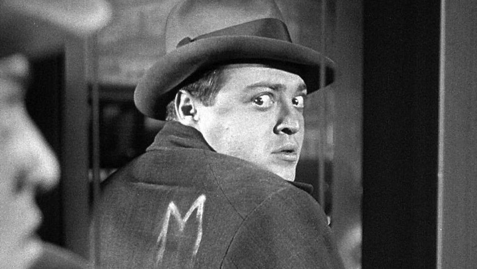 Filmstill aus M – EINE STADT SUCHT EINEN MÖRDER: Eine Mann mit Hut betrachtet im Spiegel seinen Rücken und sieht, dass auf seine Jacke in weißer Farbe der Buchstabe "M" gemalt wurde.