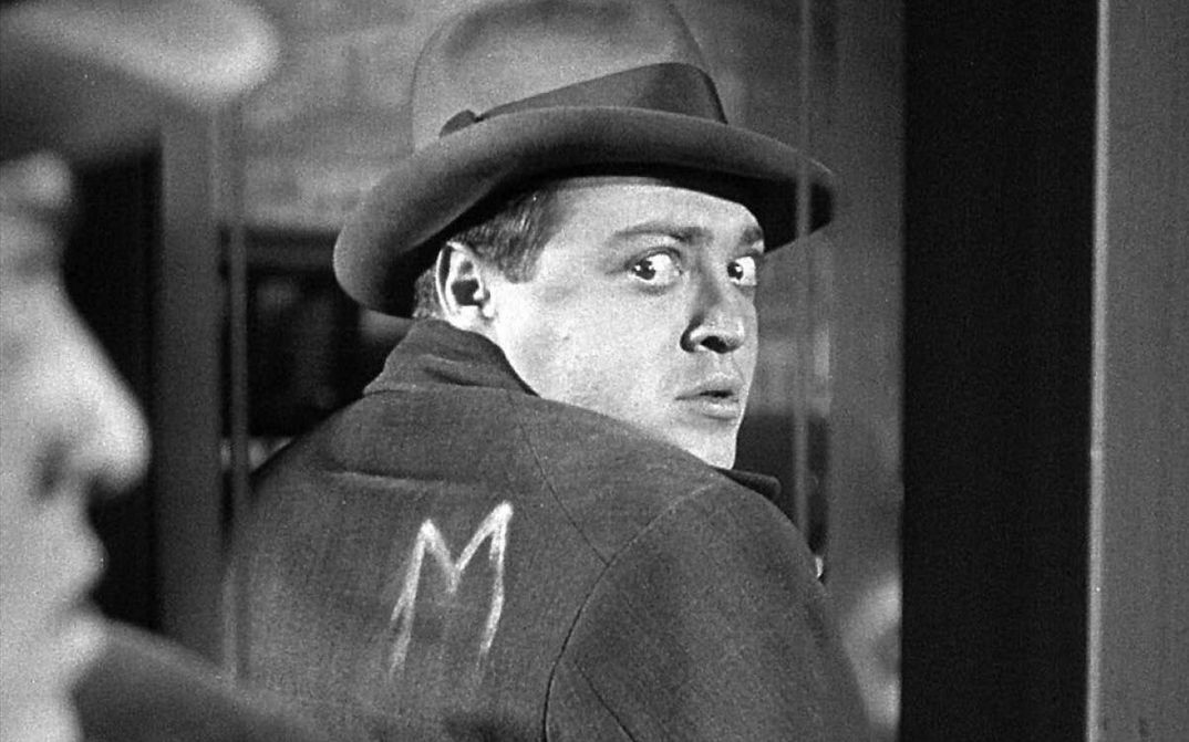 Filmstill aus M – EINE STADT SUCHT EINEN MÖRDER: Eine Mann mit Hut betrachtet im Spiegel seinen Rücken und sieht, dass auf seine Jacke in weißer Farbe der Buchstabe "M" gemalt wurde.