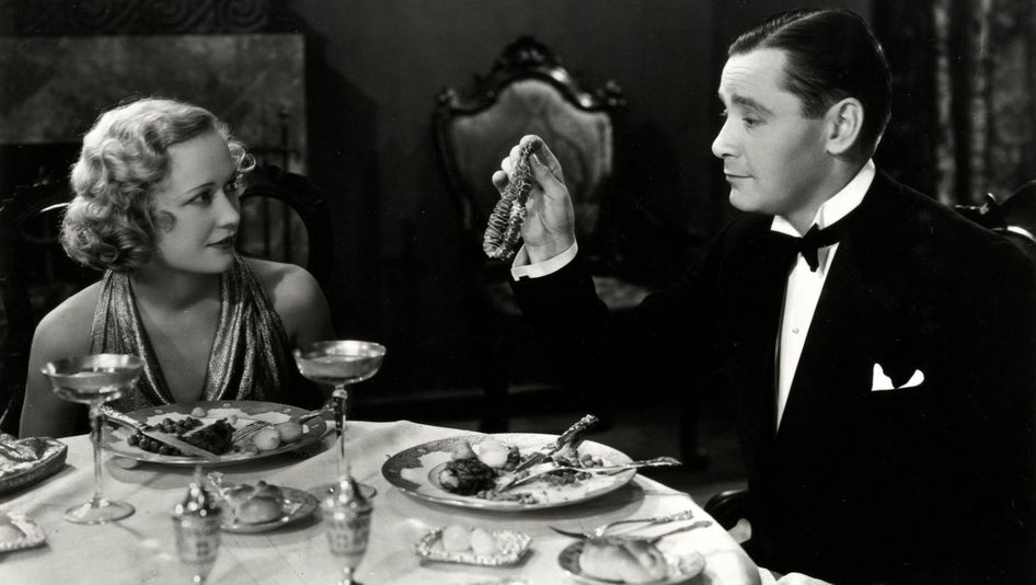 Filmstill aus TROUBLE IN PARADISE: Ein Mann und eine Frau, beide elegant gekleidet, sitzen an einem gedeckten Tisch. Er präsentiert ihr eine Kette.