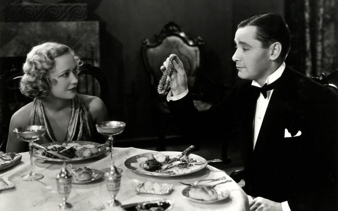 Film still from TROUBLE IN PARADISE: A man and a woman, both elegantly dressed, sit at a table. He presents her with a necklace.